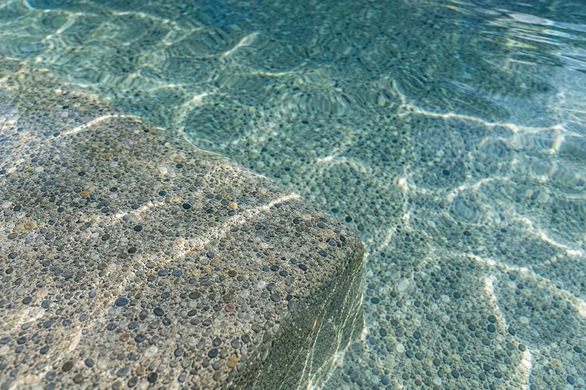 Rounded pebble aggregate set in a cement pool finish, seen through clear shallow water at a step edge