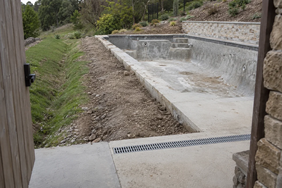 View through a gate toward an unfinished pool, hillside retaining wall, grass swale and foreground channel drain